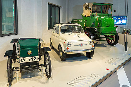 Auf einem niedrigen Podest stehen drei historische Elektro-Fahrzeuge. Von links nach rechts: ein grüner offener Wagen, ähnlich einer Seifenkiste, ein weißer Fiat 500 und ein grüner LKW mit offener Ladefläche. 