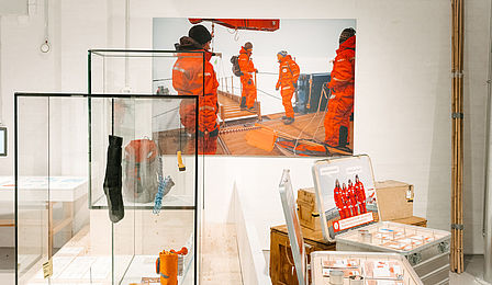 Notes and pens are laid out on aluminum boxes. Equipment, including gloves, a signal mirror and a bivouac tent, are presented in a display case next to them. 