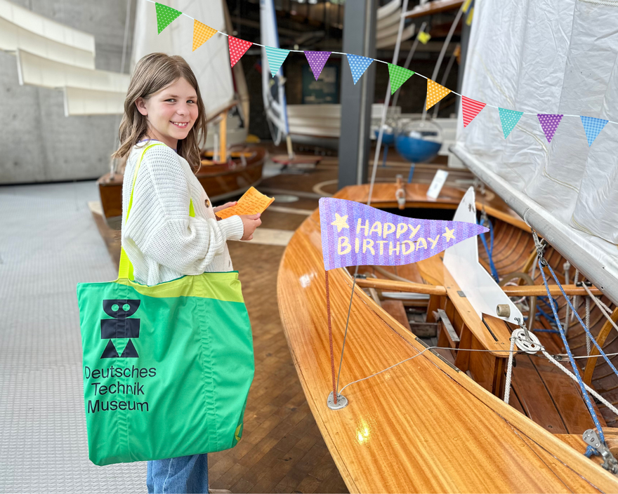 Ein Mädchen steht lachend mit einer großen grünen Museumstasche in der Schifffahrtsausstellung. In der Hand hält sie einen Brief. Das Bild wurde nachträglich mit bunten Wimpeln und einer Geburtstagsfahne dekoriert.