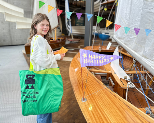 Ein Mädchen steht lachend mit einer großen grünen Museumstasche in der Schifffahrtsausstellung. In der Hand hält sie einen Brief. Das Bild wurde nachträglich mit bunten Wimpeln und einer Geburtstagsfahne dekoriert.