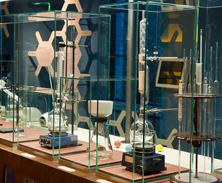There are several display cases on a table. They contain various instruments from a chemistry laboratory, including pipettes, a Liebig condenser (used for distillation), a centrifuge, and a microscope.