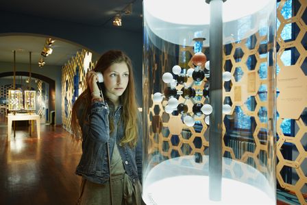 Eine Besucherin hält sich den Hörer einer Audiostation ans Ohr. Dabei sieht sie auf ein Molekül-Modell aus schwarzen, weißen und roten Kugeln.