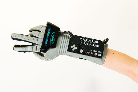 Ein Arm ragt von rechts in das Bild hinein. Hand und Unterarm sind von einem grauen Handschuh umhüllt. Ein schwarzes Interface mit vielen Knöpfen ist auf dem Handschuh befestigt.