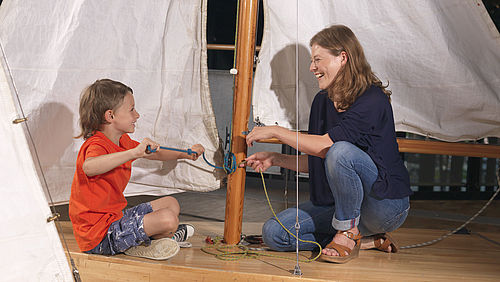 Eine Frau und ein Junge sitzen sich vor einem Segelmast mit aufgespannten Segeln gegenüber. Beide haben Seile in der Hand und machen Knoten. 
