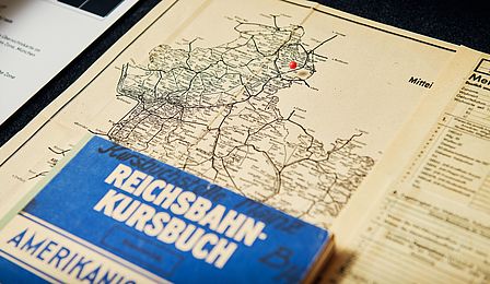Eine Landkarte mit Bahn-Streckennetzen, zu erkennen sind die Städte Frankfurt und Wiesbaden. Darüber liegt ein blaues Buch mit der Aufschrift "Reichsbahn Kursbuch".