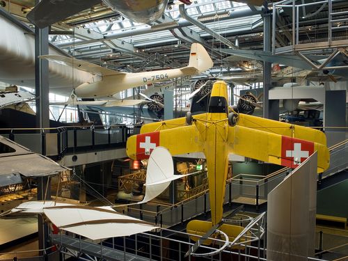 In der zentralen Treppengalerie im Neubau des Deutschen Technikmuseums hängt senkrecht ein gelber Doppeldecker. Die Tragflächen zeigen jeweils ein weißes Kreuz auf rotem Grund. Im Vorder- und Hintergrund befinden sich weitere Flugzeuge. 