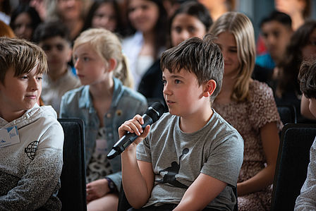Ein Junge aus dem Publikum ist seitlich zu sehen und spricht in ein Mikrofon. Im Hintergrund sitzen weitere Kinder als Teilnehmende der SchülerInnen-Konferenz.