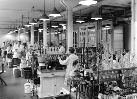 Ein Schwarzweiß-Foto zeigt Menschen in weißen Kitteln, die in einem Labor an chemischen Instrumenten arbeiten. Es ist die Pflanzenschutzforschung im Hauptlaboratorium der Schering AG im Jahr 1955.