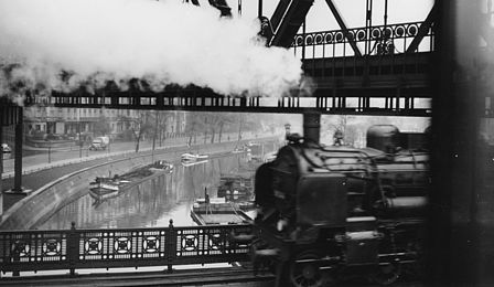 Schwarzweiß-Foto einer in Dampfwolken gehüllten Lokomotive, die auf einer Brücke einen Kanal überquert.
