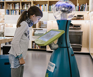 Ein Mädchen steht vor einem Roboter mit rundem Kopf, großen Augen, kegelförmigem Körper und Touchpad.