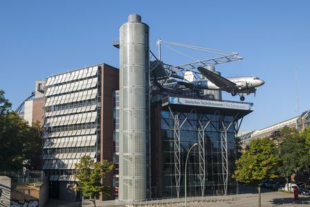 Ein modernes Neubaugebäude mit Glasfassade. An einer Stahlkonstruktion auf dem Dach hängt ein Flugzeug.