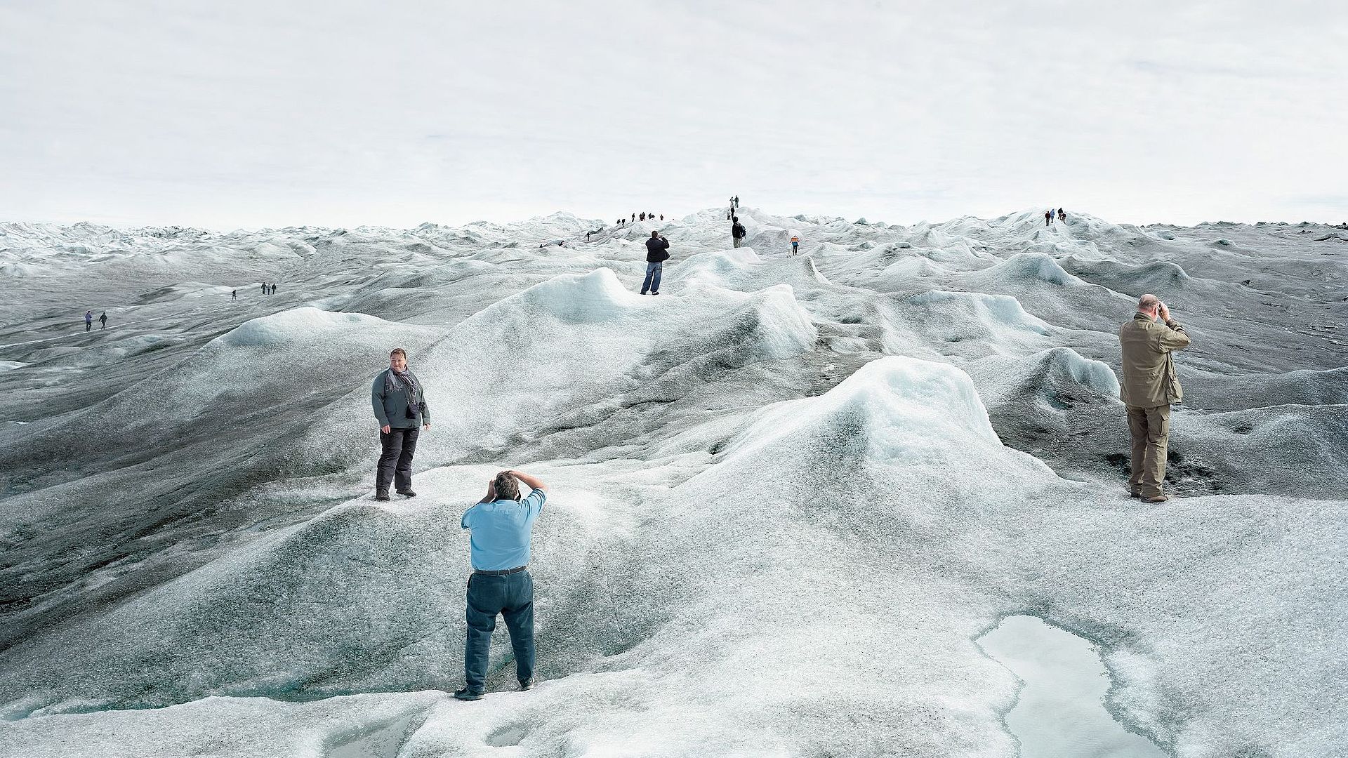 Eine Gruppe von Touristinnen und Touristen steht auf einer schmelzenden Eislandschaft und posiert an verschiedenen Punkten des Bildes für Fotos. An vielen Stellen der Eislandschaft hat sich schwarzer Ruß abgesetzt, das Eis ist dort sehr dunkel. Im Vordergrund ist eine große Pfütze aus Schmelzwasser.