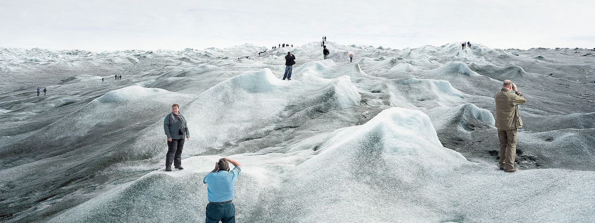 Eine Gruppe von Touristinnen und Touristen steht auf einer schmelzenden Eislandschaft und posiert an verschiedenen Punkten des Bildes für Fotos. An vielen Stellen der Eislandschaft hat sich schwarzer Ruß abgesetzt, das Eis ist dort sehr dunkel. Im Vordergrund ist eine große Pfütze aus Schmelzwasser.