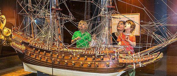 There are three children standing on a platform behind a display case, looking at a large, detailed model ship. A woman stands behind them.