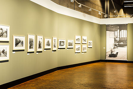 Das Bild zeigt eine Detailaufnahme der Ausstellung. An der grau-grünen Wand hängen links mehrere kleine Schwarz-Weiß-Fotos nebeneinander, hinten recht hängt ein großes Schwarz-Weiß-Foto.