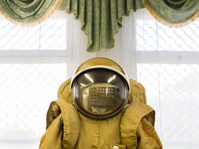 This image is a central view of a cosmonaut’s suit from the waist upwards and includes a spherical helmet with a large glass visor. In the background, an ornamental hanging with a gold fringe is suspended above a window.