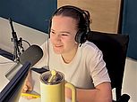 Eine junge Person mit braunen Haaren sitzt in einem Tonstudio vor einem Mikrofon und lächelt. Die person hält ein Smartphone mit einer gelben Schutzhülle in der rechten Hand.