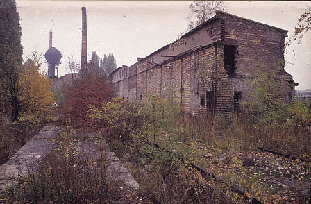 Eine Ruine eines länglichen Gebäudes in einem Park ist zu sehen, dahinter erkennt man einen Wasserturm und einen Schornstein.