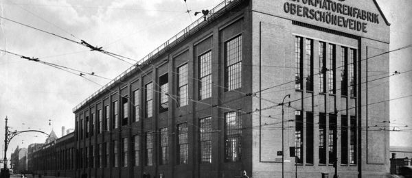 Schwarzweiß-Foto eines großen Fabrikhallengebäudes mit großen Fenstern und einem gewölbten Dach. An der Fassade ist der Schriftzug AEG zu lesen.