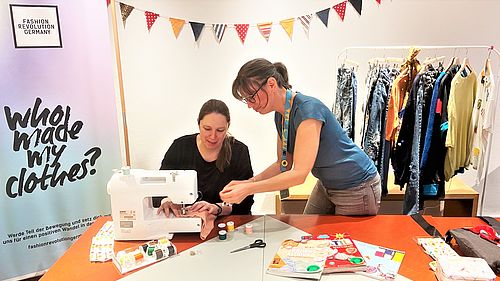 Zwei Frauen reparieren Textilien mit einer Nähmaschine. Im Hintergrund ein Kleiderständer mit verschiedenen Textilien.