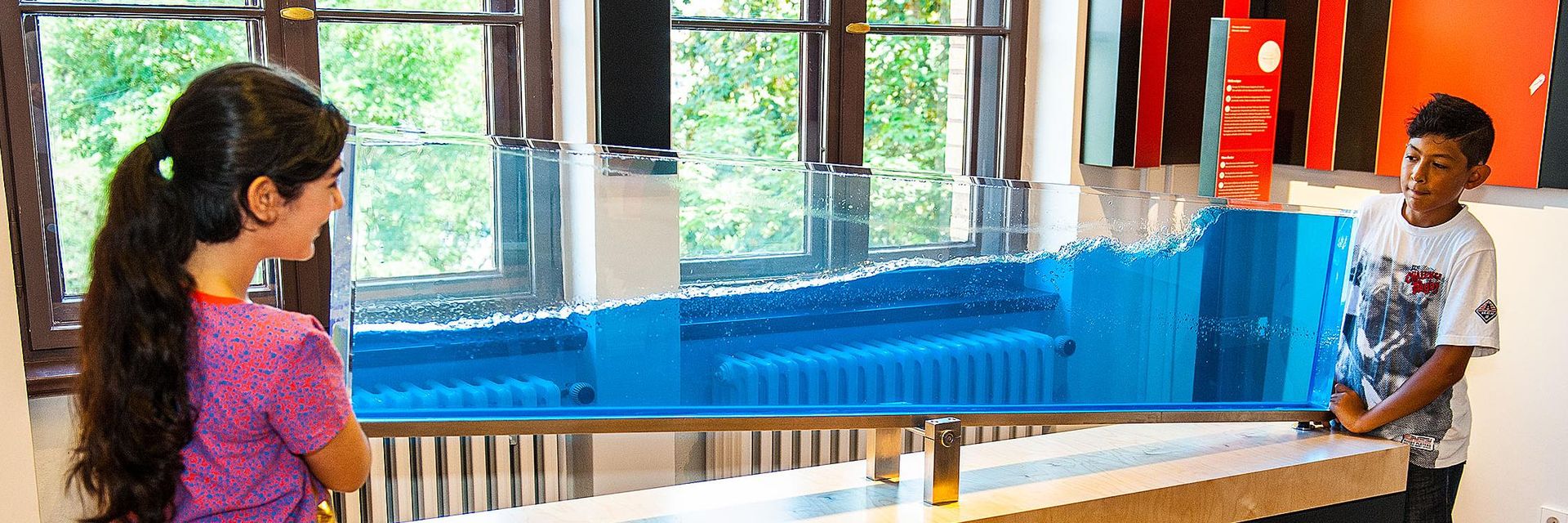 Auf einem schmalen Tisch ist transparentes Becken auf einer Kippvorrichtung befestigt. Im Becken befindet sich eine blau durchscheinende Flüssigkeit, die etwas mehr als die Hälfte des Beckens füllt. Zwei Kinder stehen an den gegenüberliegenden Enden des Tisches und kippen das Becken. An der Oberfläche der Flüssigkeit sind Wellen zu sehen.
