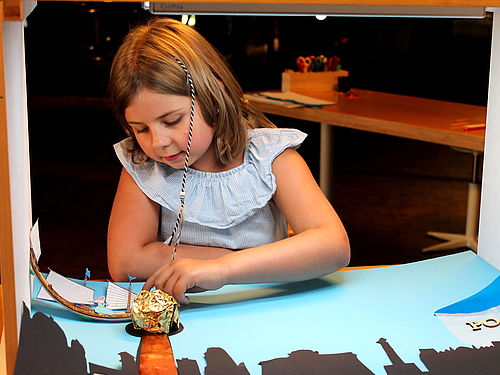 Ein Mädchen sitzt an einem Tisch, auf dem eine Kiste mit zwei offenen Seiten steht. In der Kiste liegen eine ausgeschnittene Stadtsilhouette und ein Schiff aus Papier auf einem blauen Hintergrund. Das Mädchen verschiebt das Schiff mit der linken Hand.