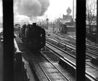 Schwarzweiß-Foto einer Dampflokomotive, die auf einer Schiene ins Bild fährt. Im Hintergrund sind ein Wasserturm und ein Lokschuppen zu erkennen.