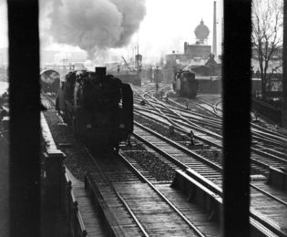 Schwarzweiß-Foto einer Dampflokomotive, die auf einer Schiene ins Bild fährt. Im Hintergrund sind ein Wasserturm und ein Lokschuppen zu erkennen.