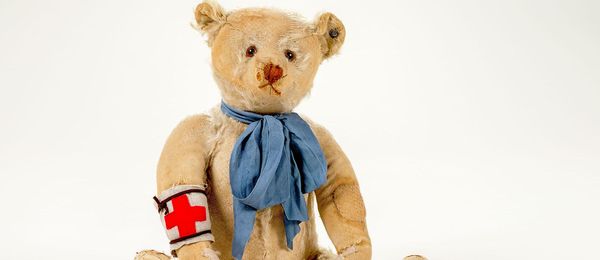 The picture shows a beige teddy bear with obviously broken and worn parts. He wears a blue bow around his neck and a white armband with a red cross.
