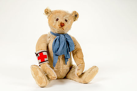 Zu sehen ist ein beiger Teddybär mit offensichtlich kaputten und abgewetzten Stellen. Er trägt eine blaue Schleife um den Hals und eine weiße Armbinde mit rotem Kreuz.