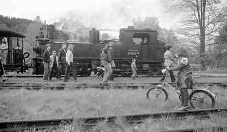 Adults and children walk across overgrown railway lines. Behind them a steam locomotive with carriages is visible. A girl stands beside her bike and is looking towards the stationary steam locomotive on which the passers-by have just arrived.