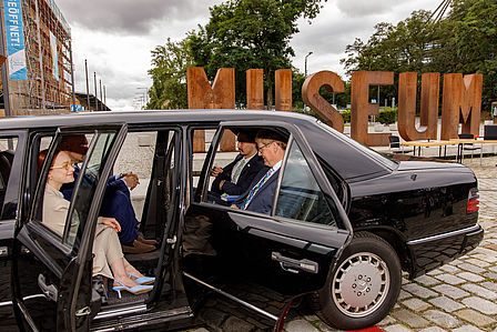 Schwarze Limousine, im Inneren sitzen drei Männer und eine Frau. Die Türen des Fahrzeugs sind geöffnet.