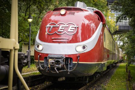 Ein roter Zug mit geschwungener Verzierung steht auf einem begrünten Gleisabschnitt.