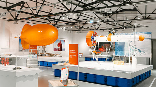 Blick in die Ausstellung. Links ein orangefarbener Fesselballon, rechts eine längliche Messsonde. Die Ausstellungseinheiten stehen auf "Schollen".