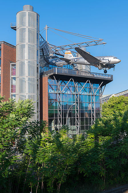 Ein modernes Neubaugebäude mit Glasfassade. An einer Stahlkonstruktion auf dem Dach hängt ein Flugzeug. Unten ist das Grün von Bäumen zu sehen.