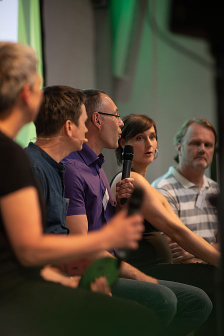 Fünf Menschen befinden sich auf der Bühne, mittig spricht eine Person ins Mikrofon. Die anderen Personen schauen ihn an.