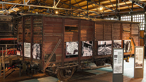 View of an old, brown wooden boxcar. Black-and-white photographs of concentration camps are displayed on the boxcar’s sides. 