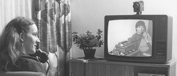 Schwarz-Weiß-Foto: Eine Frau sitzt in einem Wohnzimmer und telefoniert. Auf dem Fernseher ist das Video-Bild ihrer Gesprächspartnerin zu sehen.