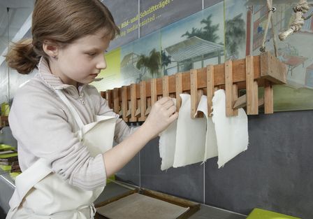 Ein Mädchen befestigt noch feuchtes, selbstgeschöpftes Papier an einer Hängevorrichtung aus Holz zum Trocknen. Das Mädchen trägt eine weiße Schürze, neben ihm liegt ein Schöpfsieb.