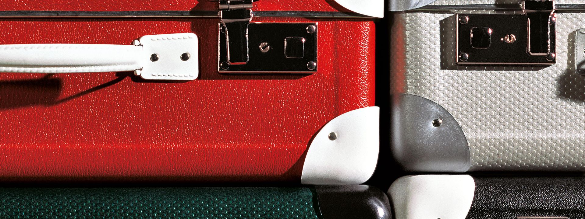 Close-up of four suitcases stacked on top of one another. One suitcase is red with white corners; one is grey with silver corners; one is black with white corners; and one is green with black corners.