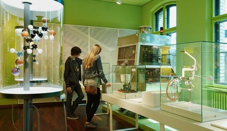 Two museum visitors look at a series of machines and instruments for turning chemical substances into tablets, pills, powders, and ointments. Next to them is a model of a penicillin molecule.