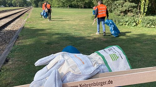Im Vordergrund Handschuhe und eine Holzzange mt der Aufschrift "www.kehrenbürger.de", im Hintergrund ein Park, in dem Menschen mit orangefarbener Weste und blauen Müllbeuteln umhergehen.