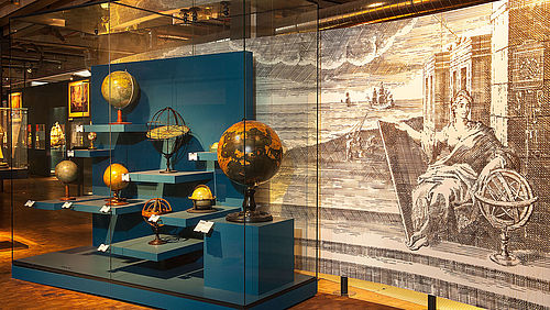 Vor einem großen, schwarz-weißen Wandbild stehen in einer Vitrine mit blauen Sockeln verschiedene historische Globen.