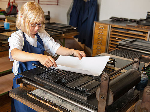 Ein Mädchen steht vor einer kleinen Druckerpresse und legt vorsichtig ein Blatt Papier auf einen gesetzten Text. Sie trägt eine Schürze und hält das Blatt behutsam mit einer Hand an jeder Seite fest.