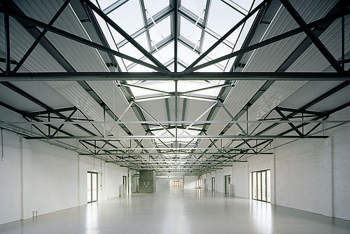 Blick in eine Lagerhalle, die offensichtlich neu restauriert wurde: Die Böden und Wände glänzen weiß, die Stahlkonstruktion der Decken geben den Blick auf Glasgauben frei.
