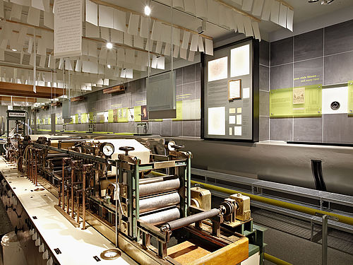 Blick in die Ausstellung Papiertechnik. Im Vordergrund steht eine mehrere Meter lange Maschine, von der Decke hängen Papierbögen, an der Wand sind Bilder und Texte angebracht.