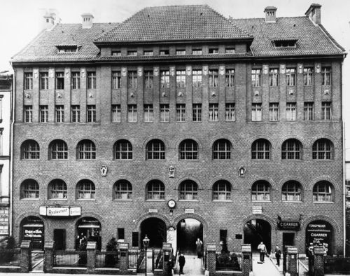 Schwarzweiß-Foto: Frontalansicht eines großen, vierstöckigen Backsteingebäudes. Im Erdgeschoss führen große Torbögen ins Innere.