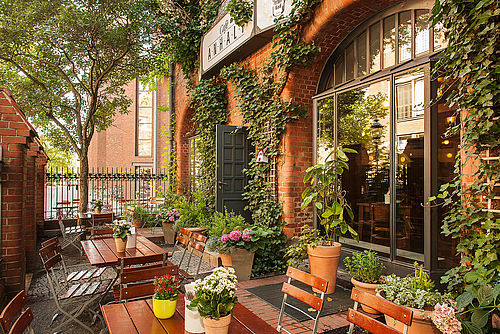 Blick auf die begrünte Terrasse eines Restaurants, mit einladend dekorierten Holztischen und Stühlen.