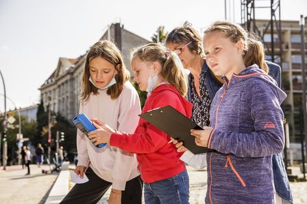 Drei Mädchen stehen auf dem Vorplatz des Museums und halten Tablets in der Hand, auf die sie konzentriert schauen.