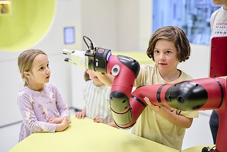 Drei Kinder stehen an einem rot-schwarzen Roboterarm. Ein kleiner Junge bewegt den Roboterarm mit seinen Händen. 